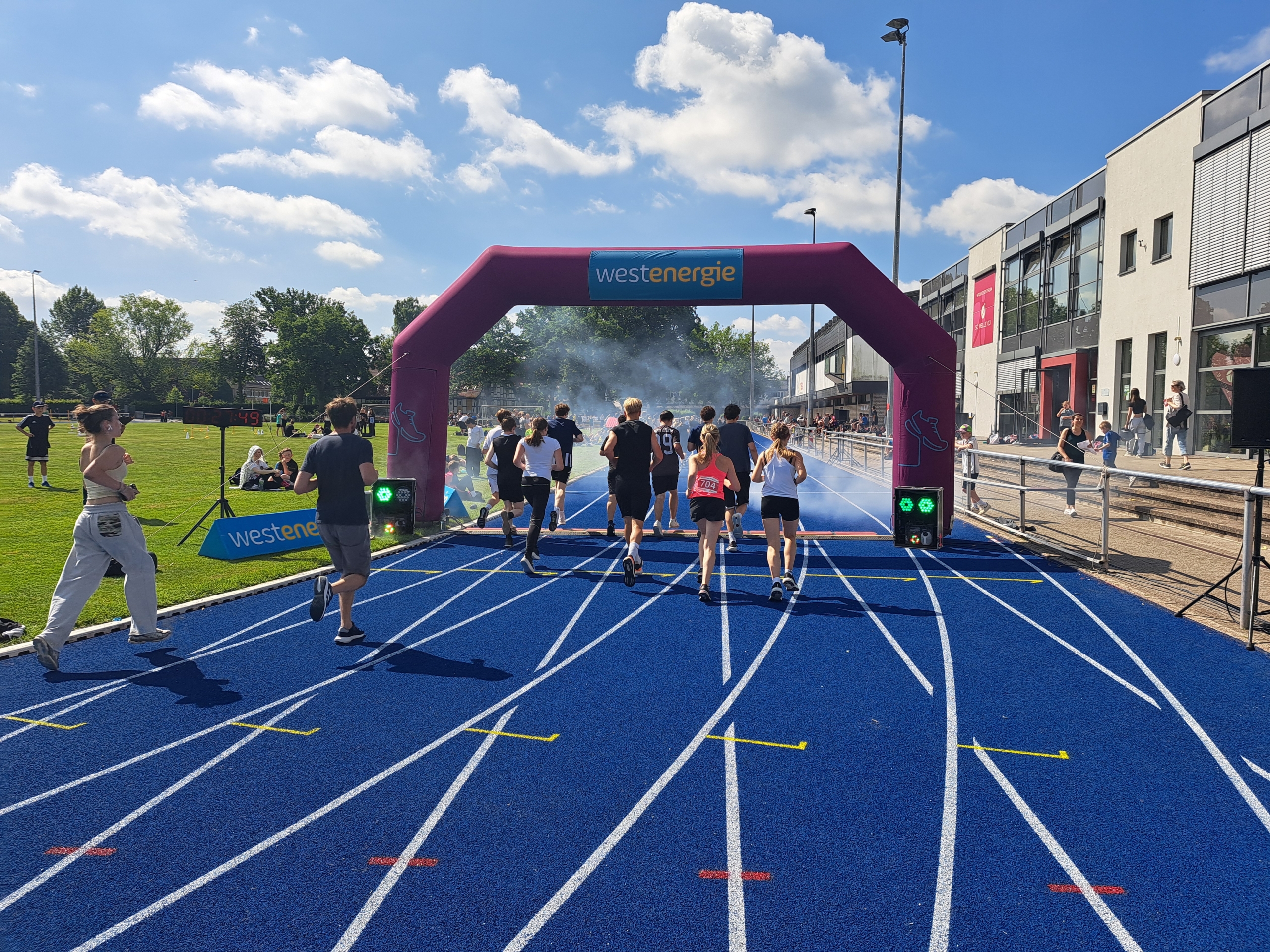 Das Bild zeigt eine Laufveranstaltung bei sonnigem Wetter auf einer blauen Tartanbahn mit weißen Linien. Mehrere Jugendliche und junge Erwachsene laufen auf den Zielbogen zu, der über der Bahn aufgestellt ist. Der aufblasbare Bogen ist lila und trägt oben ein Schild mit der Aufschrift „Westenergie“ in Blau und Gelb. Links neben der Laufbahn befindet sich eine große grüne Rasenfläche, auf der einige Zuschauerinnen und Zuschauer sitzen oder stehen. Dort ist auch eine digitale Zeitanzeige mit der Zahl 4:49 zu sehen. Rechts neben der Bahn steht ein modernes, weißes Gebäude mit großen Fenstern. Hinter dem Bogen ist leichter Nebel oder Rauch zu sehen, wahrscheinlich Teil eines Effekts für das Laufevent. Die Stimmung ist lebhaft und sportlich, der Himmel ist blau mit einigen weißen Wolken. Das Bild vermittelt eine fröhliche Atmosphäre bei einer sportlichen Veranstaltung im Freien.